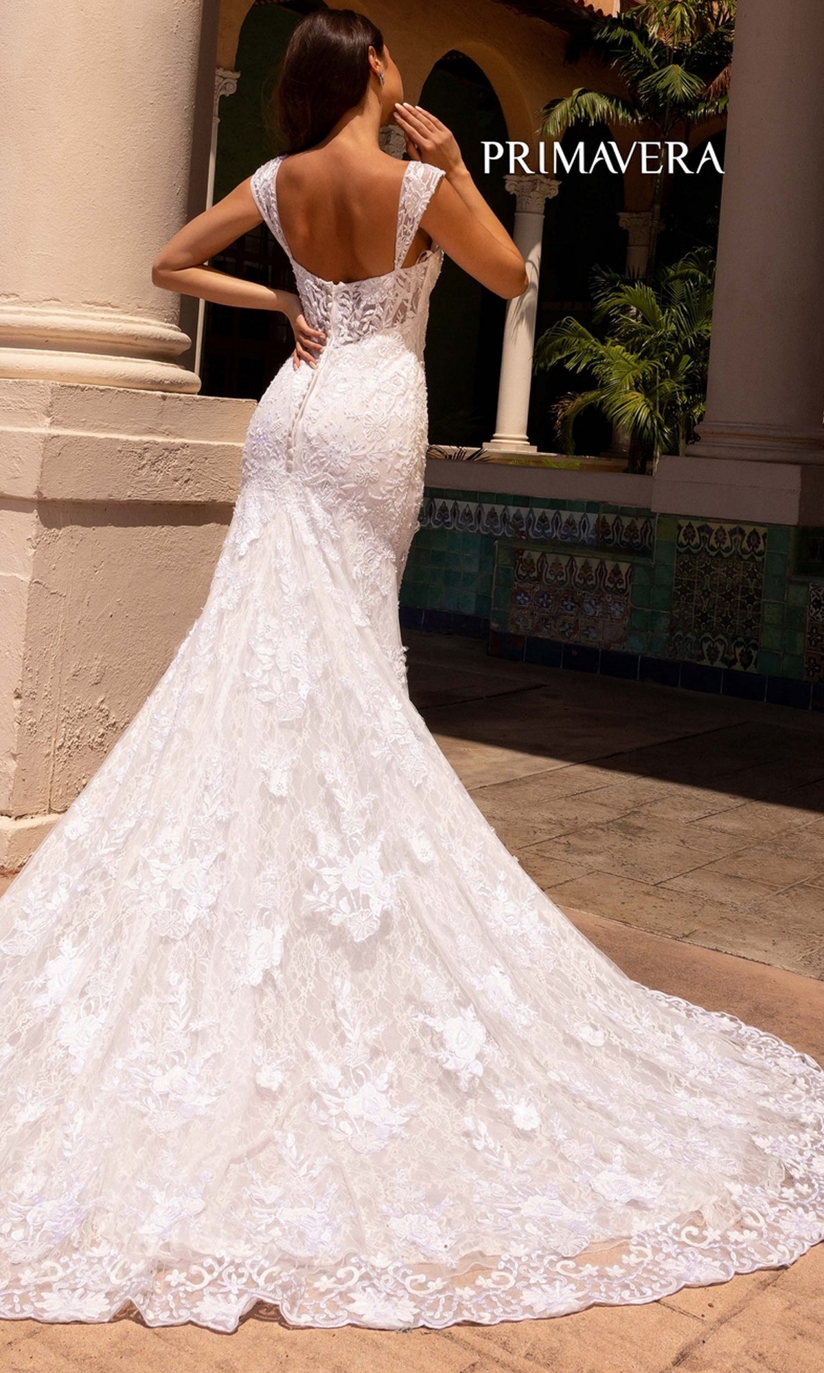 A woman stands outdoors near columns and tiled walls wearing the Long Wedding Dress 11107 by Primavera, an ivory fitted gown with cap sleeves, long train, buttoned back, and deep V-neckline.